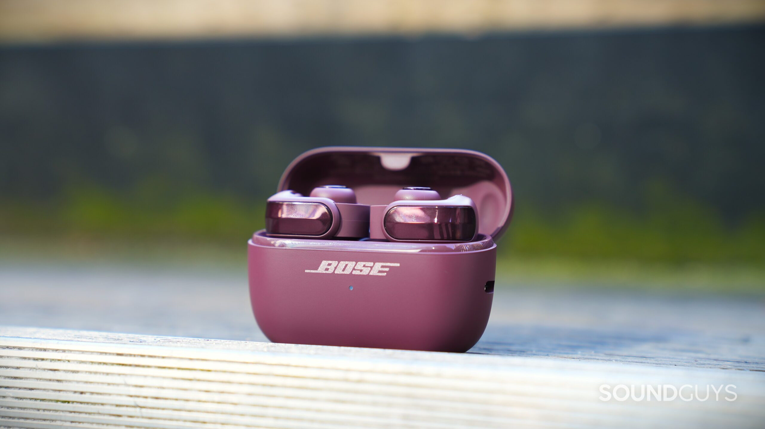 DSC09919 Cherry Chocolate Bose Ultra Open 2 earbuds in their open charging case on a wooden railing outdoors.