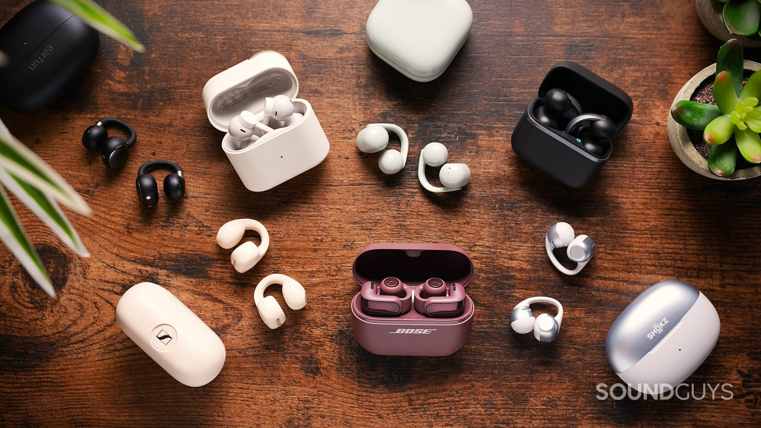 best clip-on earbuds Various clip-on open-ear earbuds and charging cases arranged on a wooden table, with burgundy Bose earbuds in an open case at the center and plants framing the scene.